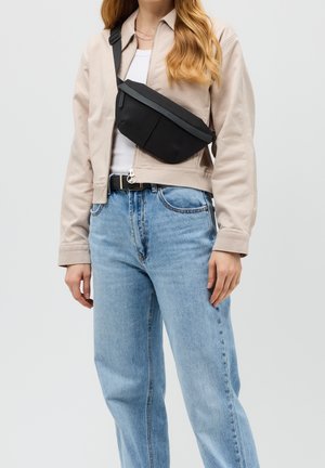 Zwarte schoudertas met een gestructureerde afwerking, gebogen vorm en verstelbare schouderband, gedragen over een lichtgekleurde jas en jeans.