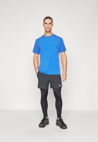 Sinine spordisärk väikeste aukudega, mustad lühikesed püksid valge Nike logo ja mustad retuusid, mustad spordijalatsid, kõik ühetaolisel taustal.