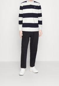 Uomo in polo a maniche lunghe a righe bianche e nere, pantaloni neri e sneakers bianche, in piedi su uno sfondo bianco uniforme.