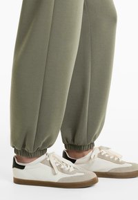 Olivengrønne sweatpants med elastiske manchetter, parret med beige sneakers med et tekstureret hvidt og sort design og cremede snørebånd.