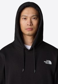 Svart hoodie i mjukt tyg, med en framficka och dragskor. På vänster bröst finns en vit logotyp för The North Face.