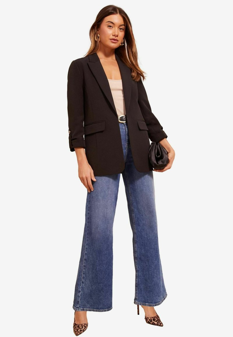 Schwarzer taillierter Blazer mit Reverskragen, kombiniert mit hochtaillierten Schlagjeans. Die Accessoires umfassen eine schwarze Clutch und hohe Schuhe im Tiermuster.