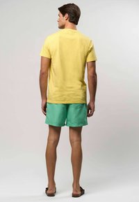 Kollane puuvillane t-särk ja rohelised lühikesed püksid tagataskuga, kantud pruuni sandaalidega, nähtud tagant lihtsa tausta ees.