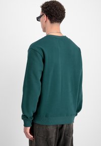 Sudadera verde de cuello redondo con puños y dobladillo acanalados. Tejido suave, con una pequeña etiqueta rectangular en la parte posterior cerca del escote.