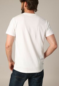 Hombre con barba vistiendo una camiseta blanca de manga corta y jeans azul oscuro, de pie con una mano en la cadera, de espaldas a la cámara.