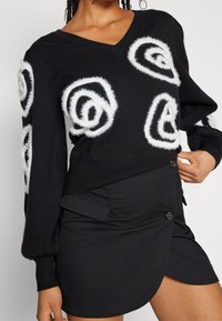 Black sweater with V-neck, featuring white fuzzy circular patterns. Paired with a black skirt that has a button detail and a distinct hemline.