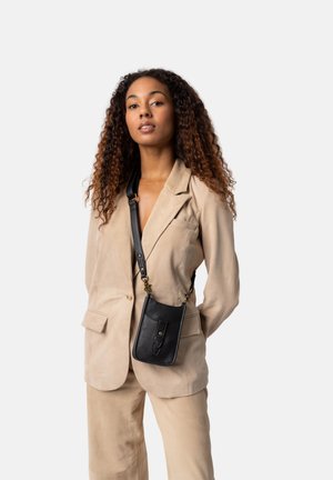 Bolso cruzado de cuero negro con textura suave, que presenta un bolsillo frontal y una correa desmontable. Llevado sobre un blazer beige.