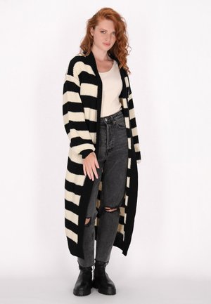Cardigan lungo a righe nere e crema con apertura frontale, lavorazione a maglia testurizzata e polsini a costine. Indossato sopra un top chiaro e abbinato a jeans a vita alta.