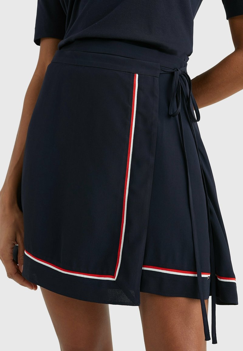 Jupe portefeuille bleu marine avec bordure rouge et blanche portée par une personne, nouée avec un nœud sur le côté, assortie d'un haut bleu marine.