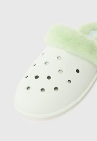 Vita slip-on träskor med cirkulära ventilationshål och en ljusgrön, fluffig foder runt öppningen för ökad komfort.