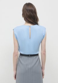 Donna con lunghi capelli castani che indossa un top senza maniche azzurro chiaro e una gonna grigia a righe, vista di spalle contro uno sfondo bianco.