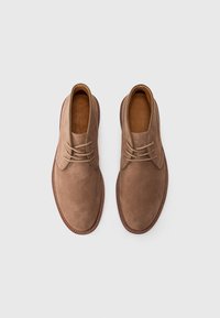 Een paar lichtbruine suede veterschoenen met ronde neuzen en gestikte bruine zolen, van bovenaf bekeken op een witte achtergrond.