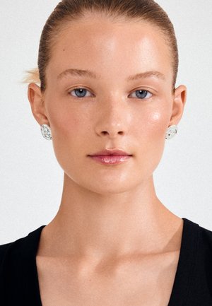 Nuori nainen, jolla on taakse sidottu tukka, siniset silmät, hopeiset kukkakorvakorut ja musta toppi, katsoo eteenpäin neutraalilla ilmeellä.