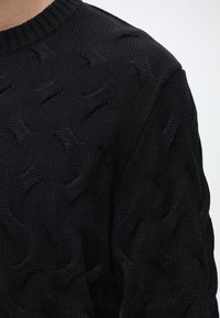Gros plan d'un pull noir à torsades présentant des motifs texturés et torsadés sur l'épaule et le haut du bras.