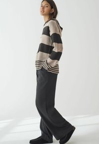 Haut à capuche en tricot rayé beige et noir, arborant une capuche et une poche. Associé à un pantalon ample foncé et des chaussures noires à enfiler.