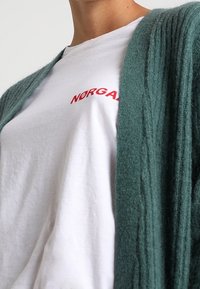 Cardigan verde a coste sovrapposto a una t-shirt bianca con testo rosso. Il cardigan ha una texture morbida e un design aperto sul davanti.