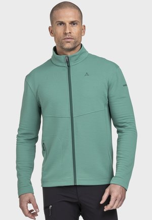 Mann trägt eine hellgrüne Jacke mit Reißverschluss und hohem Kragen sowie schwarze Hosen, steht vor einem einfarbigen Hintergrund.