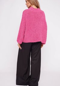 Fuzzy, bright pink cardigan with a relaxed fit, long sleeves, and a high collar. Worn over loose black trousers, showing texture and color contrast.