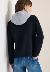 Femme portant un sweat à capuche noir avec une capuche grise et un jean bleu, debout de profil avec une main dans la poche arrière, devant un mur clair.