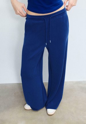 Osoba ubrana w luźne niebieskie spodnie dresowe z szerokim ściągaczem i białe wsuwane buty, stojąca na beżowej podłodze przy białej ścianie.