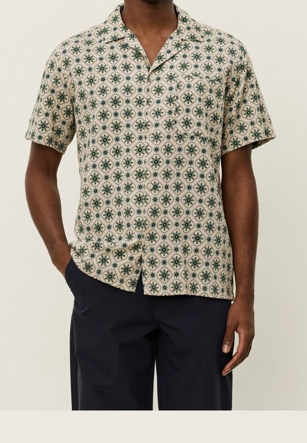 Homme portant une chemise beige à manches courtes avec un motif circulaire vert et un pantalon bleu marine, debout avec une main dans la poche.