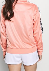 Pink satin tracksuit jacket with white and black side stripes, ribbed cuffs, and hem. Paired with white shorts, smooth texture.