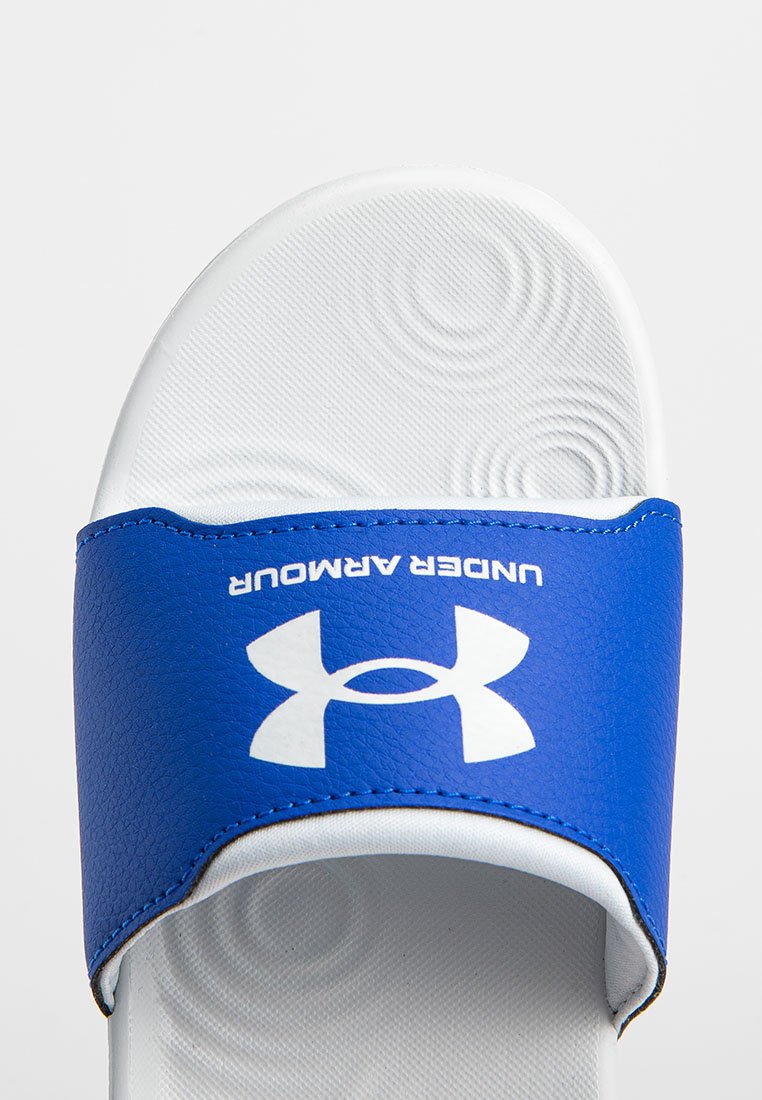 Chancla azul con suela texturizada blanca. Presenta una amplia tira azul con el logo blanco de "Under Armour", hecha de material sintético suave.