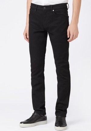 Jean noir slim porté par une personne debout avec des baskets basses noires sur un fond blanc.