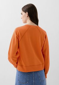 Sweatshirt orange avec un col et des poignets côtelés, présentant des manches raglan et une coupe décontractée. Associé à une jupe en denim.