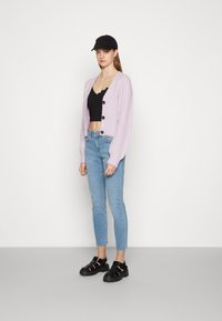 Cardigan canelado de cor roxa clara com botões pretos, usado sobre um top cropped preto, combinado com jeans skinny azuis claros e sandálias pretas.