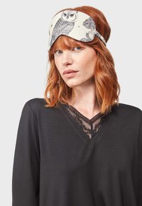 Masque de sommeil en tissu crème à motif de hiboux avec des illustrations sombres, doté d'un design contourné et d'une bande élastique ajustable.