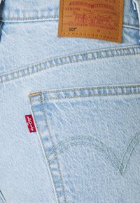 Los pantalones de mezclilla de color azul claro cuentan con una etiqueta de marca de cuero, costuras amarillas y una etiqueta roja de Levi's en el bolsillo trasero. Diseño distintivo de costura curvada.