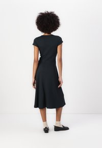 Schwarzes, kurzärmliges Kleid mit tailliertem Oberteil und ausgestellt Rock, aus weichem Stoff gefertigt. Gestylt mit schwarzen Schuhen und hellen Socken.