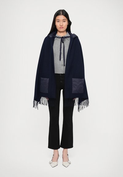 Poncho marine avec capuche et franges, doté de poches matelassées navy, superposé sur un pull gris et un pantalon évasé noir.