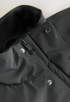 Gulerul unei jachete negre, impermeabile, cu fermoar și nasturi tip snapp; materialul are un finisaj texturat cu linii verticale subtile.