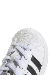 Baltās ādas sporta kurpes ar teksturētu pirksta daļu, pelēkiem akcentiem un melnām svītrām. Iezīmējas ar plakniem šņorēm un klasisku dizainu.