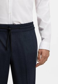 Marinebroek met een gestructureerde afwerking, elastische tailleband en verstelbare trekkoord, gecombineerd met een witte overhemd. Hand op de broek geplaatst.