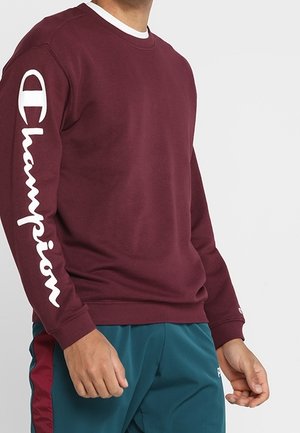 Dieprode sweatshirt met wit "Champion" logo op de linker mouw, gedragen met teal sportbroek met dieprode zijstrepen.