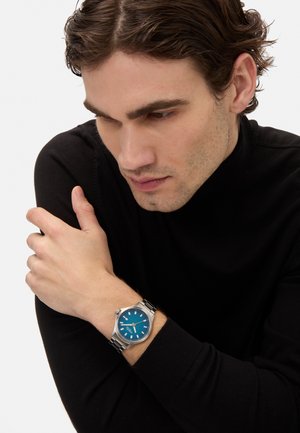 Montre bracelet en argent avec un cadran bleu, index blancs et bracelet en acier inoxydable. Le modèle porte un pull col roulé noir.