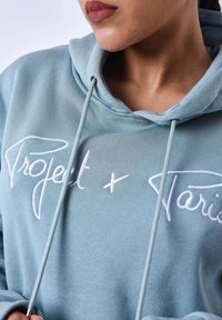 Hoodie bleu clair avec cordon de serrage, texture douce et texte brodé blanc proéminent indiquant "Projet x Paris."