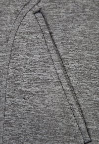 Šedý materiál mikiny s kapucí má měkký, texturovaný vzhled a obsahuje boční kapsu v kontrastní barvě a prošívané detaily podél švů.