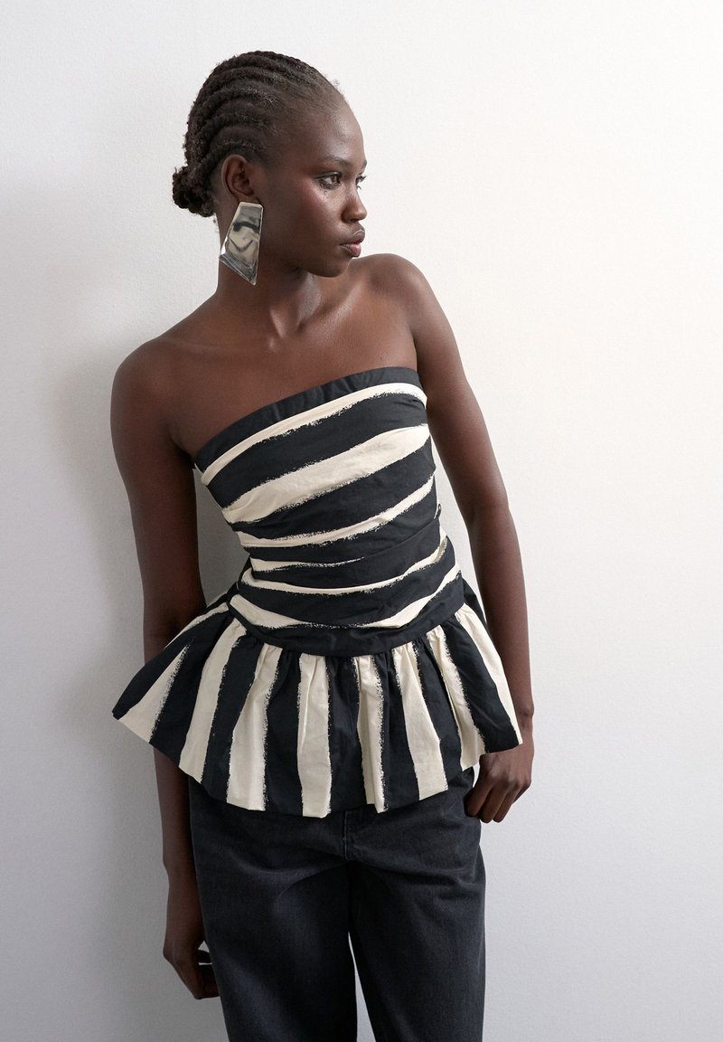 Γυναίκα με πλεγμένα μαλλιά φοράει τοπ πεπλούμ χωρίς τιράντες με μαύρες και λευκές ρίγες και μεγάλα γεωμετρικά ασημένια σκουλαρίκια, στέκεται μπροστά σε λευκό τοίχο.