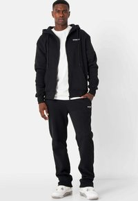 Schwarze Zip-Up-Hoodie mit Kapuze, Fronttaschen, kombiniert mit passenden schwarzen Jogginghosen. Das Model trägt ein weißes T-Shirt darunter.