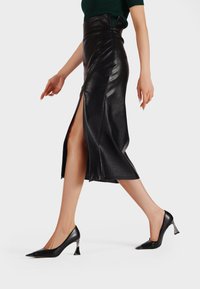 Gonna midi nera in pelle con spacco frontale, abbinata a décolleté nere a punta con un accento metallico sul tallone.