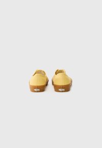 Yellow canvas slip-on shoes with rubber soles, featuring stitching details and a logo on the heel. Rear view, smooth texture.