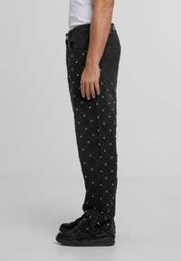 Sānskats uz personu, kas valkā melnus džinsus ar maziem sudrabotiem elementiem dimanta rakstā un melnām kedām.