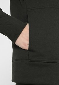 Sudadera verde oscuro con puños acanalados y un bolsillo en el frente. La tela parece suave con una textura lisa. Detalles de costura sutiles visibles.