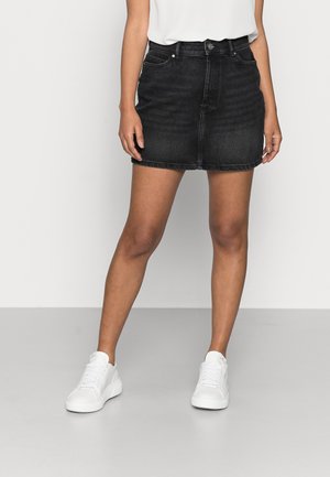 Kvinde iført en sort denim mininederdel, hvid indstukket top og hvide sneakers, stående mod en ensartet lys baggrund.