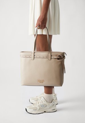 Borsa a spalla in pelle beige con doppi manici, dettagli con borchie e logo impresso sulla parte anteriore, posizionata su uno sfondo chiaro.