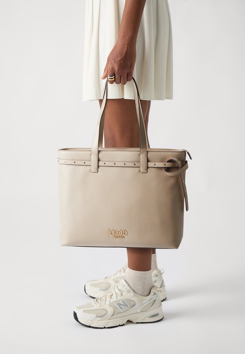 Sac fourre-tout en cuir beige avec poignées doubles, accents cloutés et logo embossé à l'avant, présenté sur un fond clair.